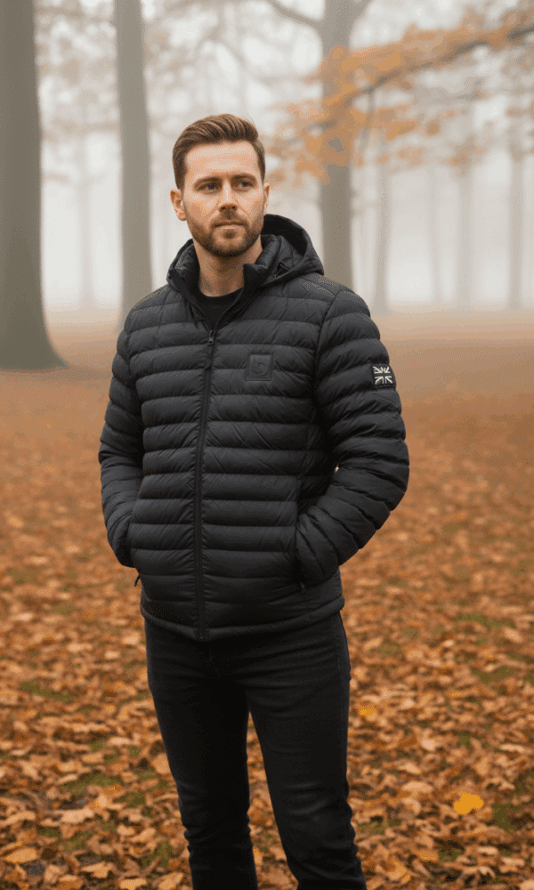 Full-body shot of a man wearing a black padded DROW Wear coat with Union Jack patch, standing in an autumn woodland.