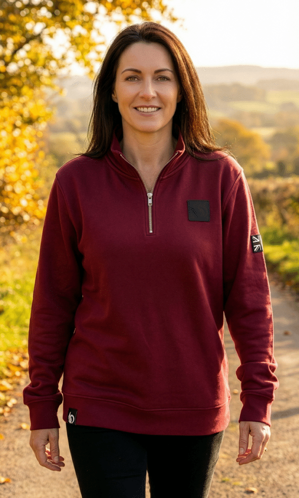 Woman wearing the DROW Wear burgundy quarter-zip jumper outdoors in autumn light, showing the British heritage Union Jack patch on the sleeve.