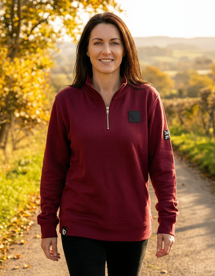 Woman wearing the DROW Wear burgundy quarter-zip jumper outdoors in autumn light, showing the British heritage Union Jack patch on the sleeve.